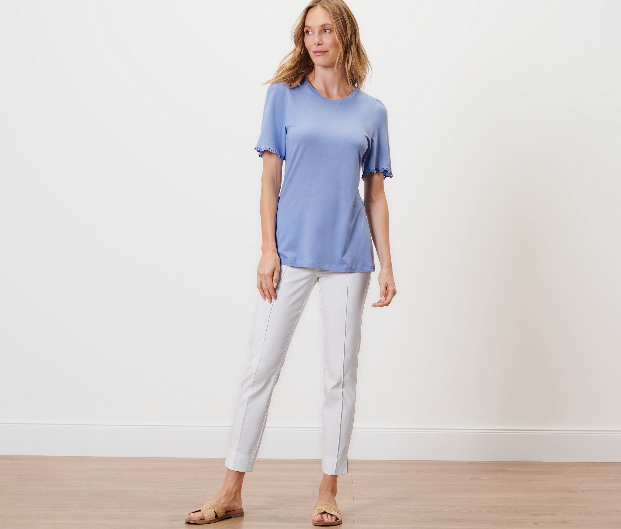 Kobieta stoi w jasno niebieskiej koszulce z marszczeniem i białych spodniach.