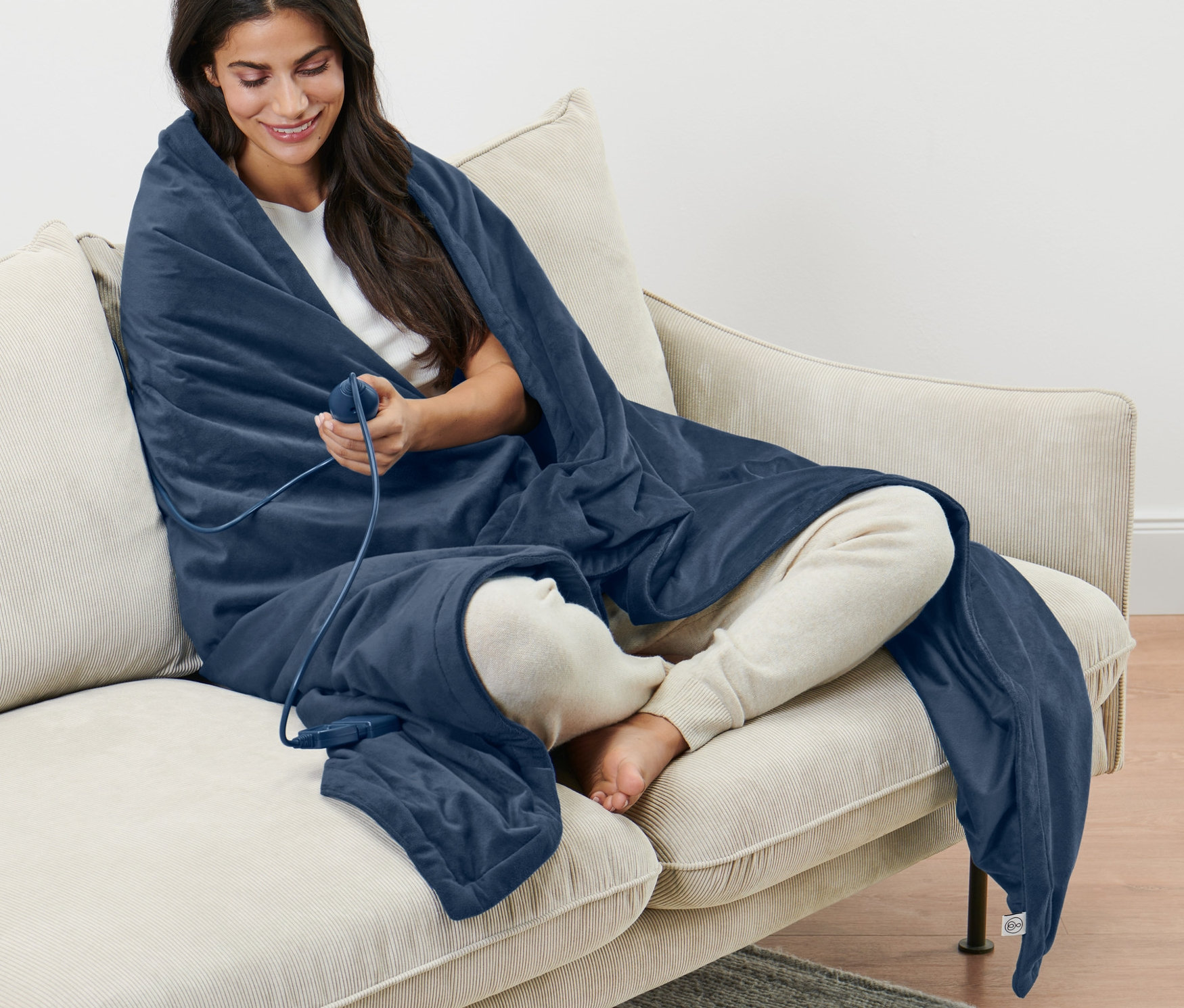 Kobieta siedzi na beżowej sofie, owinięta kocem elektrycznym.