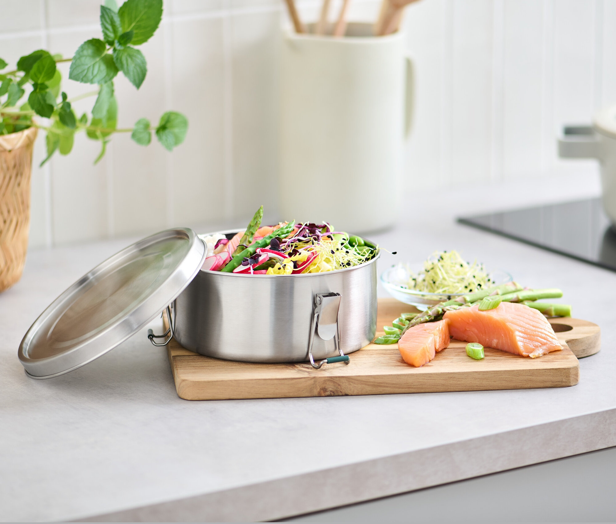 Pojemnik na lunch ze stali nierdzewnej wypełniony warzywami i sałatą obok łososia i szparagów na desce do krojenia.