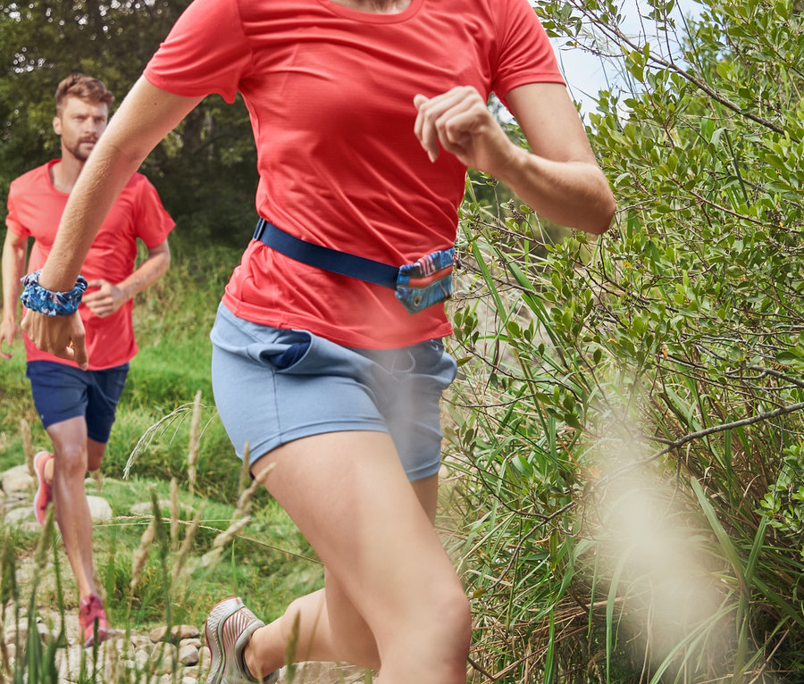 Kobieta w koralowej koszulce sportowej i granatowych legginsach sportowych o długości 3/4 biegnie po kamienistej ścieżce. W tle biegnie mężczyzna w sportowych szortach i koralowej koszulce sportowej.
