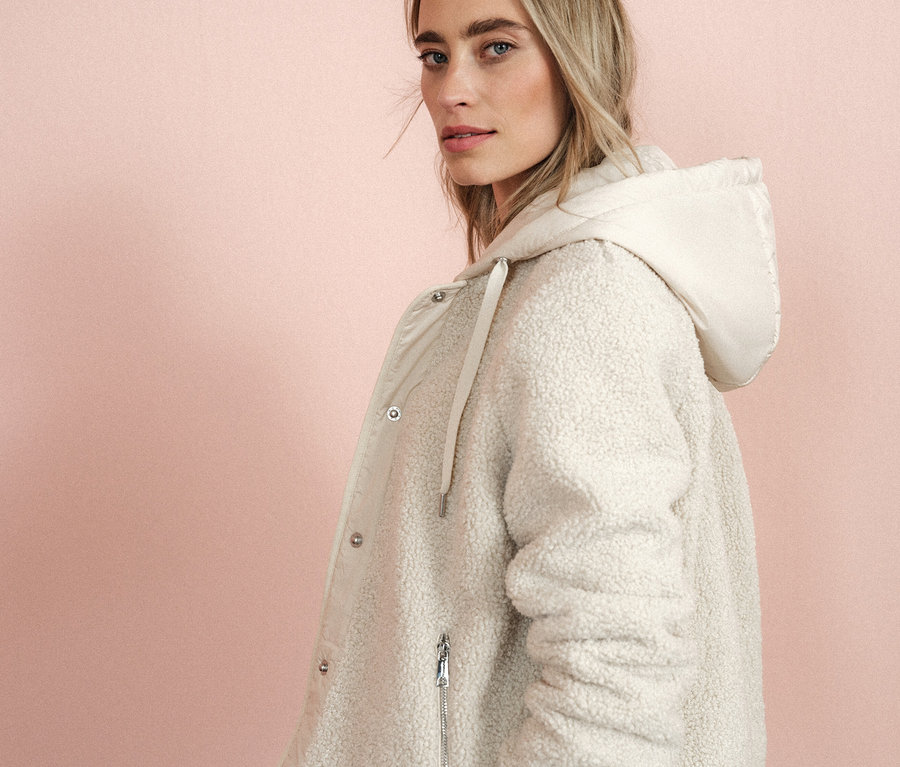 Kobieta patrzy przez ramię, ubrana w kremowy płaszcz z kapturem.