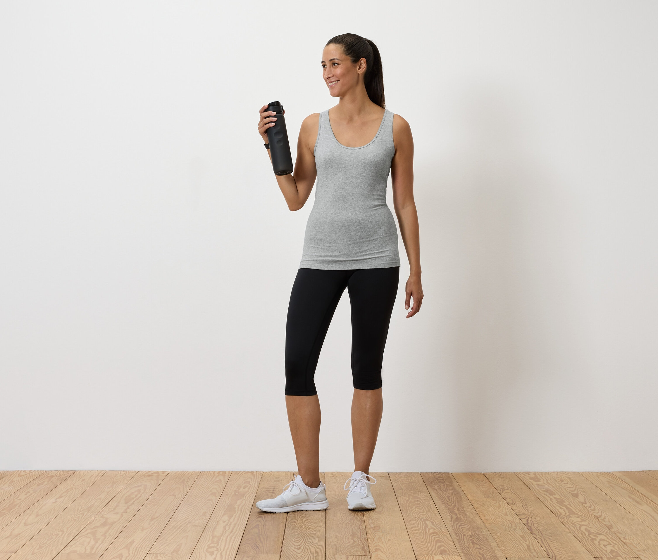 Kobieta trzyma butelkę z wodą, ma na sobie szary podkoszulek, czarne legginsy i białe tenisówki.