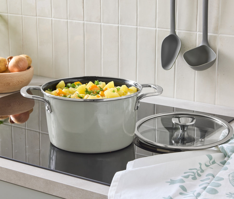 Na płycie grzewczej stoi garnek z pokrywką i warzywami. W tle miska z ziemniakami i wiszące chochle.