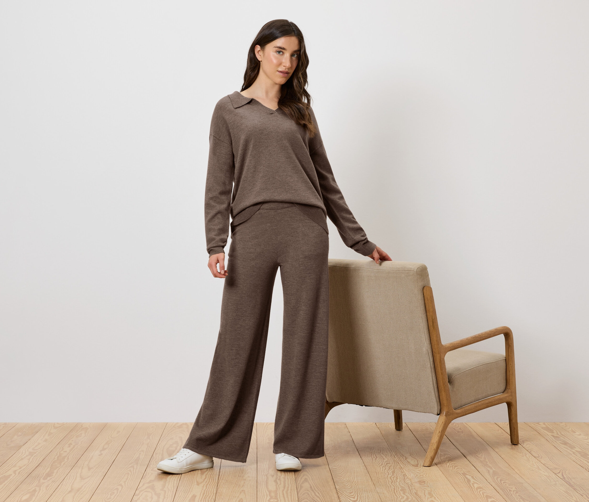 Kobieta w brązowym dzianinowym komplecie pozuje obok fotela.