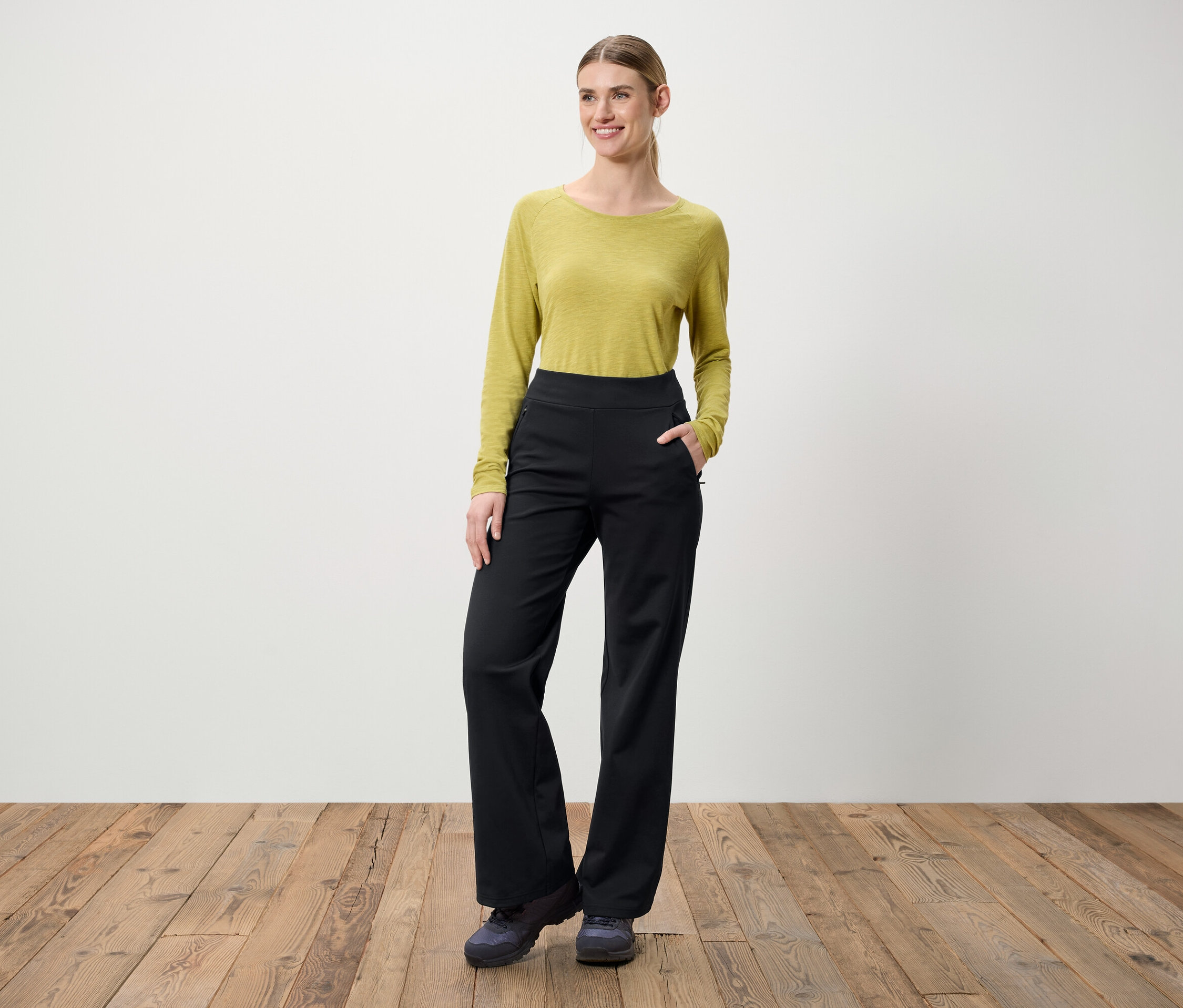 Kobieta stoi na drewnianej podłodze. Ma na sobie zieloną bluzkę z długim rękawem i czarne spodnie.