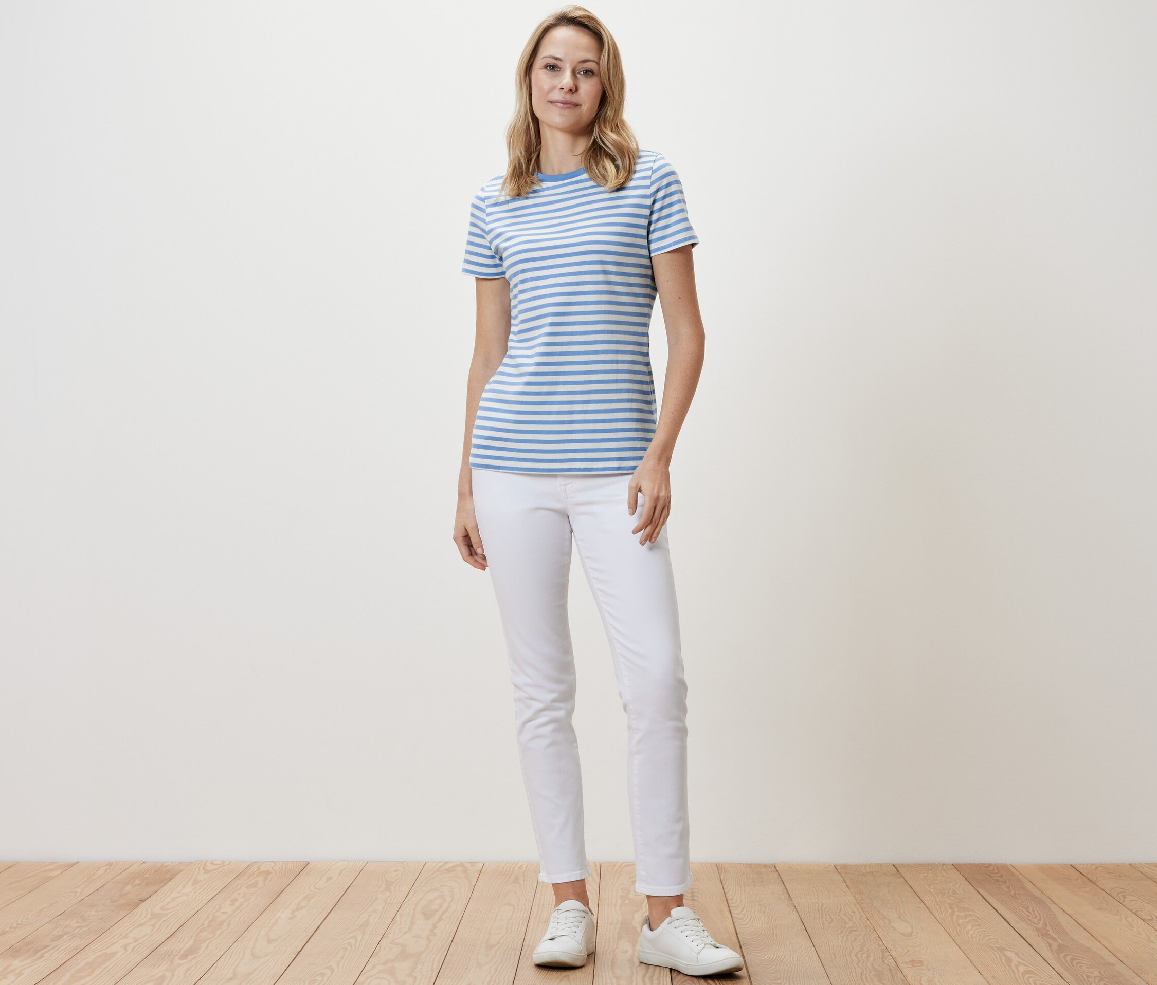 Kobieta stoi i ma na sobie koszulkę w jasnoniebiesko-białe paski, białe spodnie i białe tenisówki.