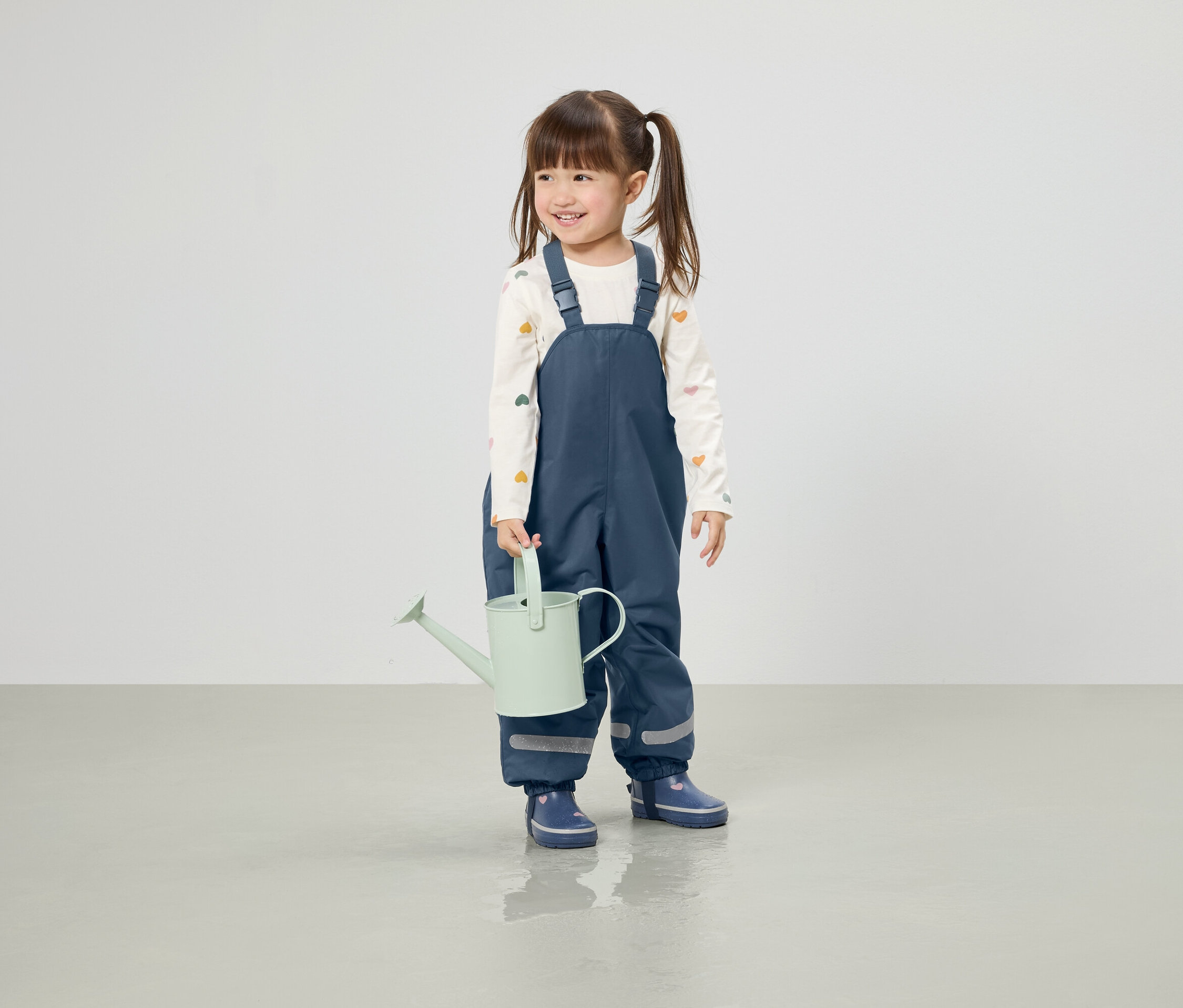 Uśmiechnięta dziewczynka w kucykach trzyma jasnozieloną konewkę, ubrana w niebieskie spodnie i kalosze.