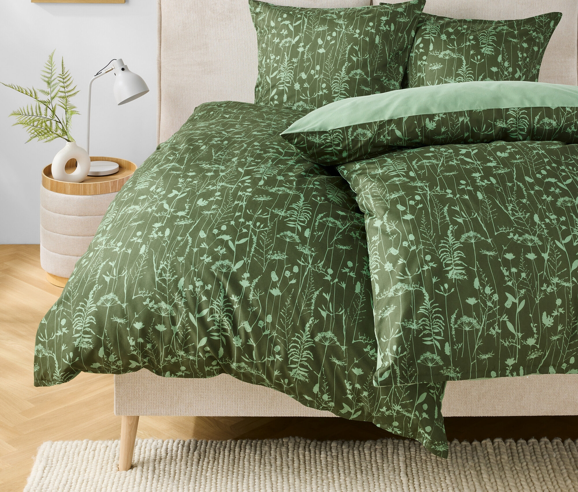 Zielona pościel w kwiatowy wzór na łóżku. Na stoliku nocnym stoi lampka i wazon z rośliną.