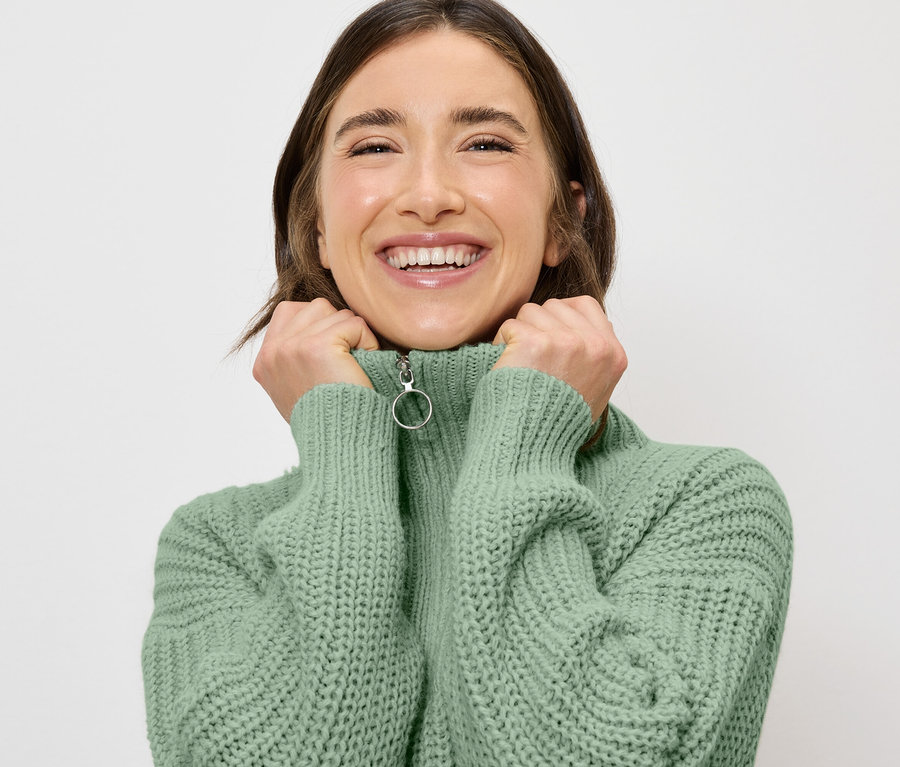 Uśmiechnięta kobieta trzyma rękoma kołnierz jasnozielonego swetra z zamkiem błyskawicznym.