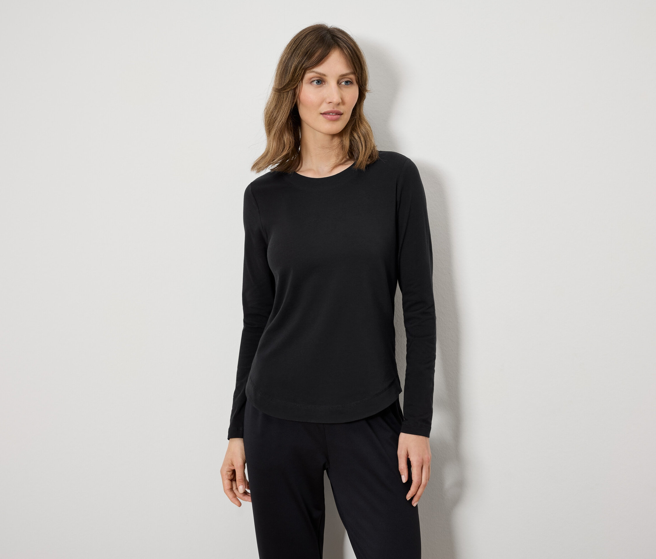 Kobieta w czarnej bluzce z długim rękawem i czarnych spodniach stoi na tle białej ściany.