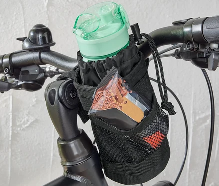 Na kierownicy roweru przymocowana jest rowerowa torebka na przekąski, w której znajduje się butelka z wodą i baton.