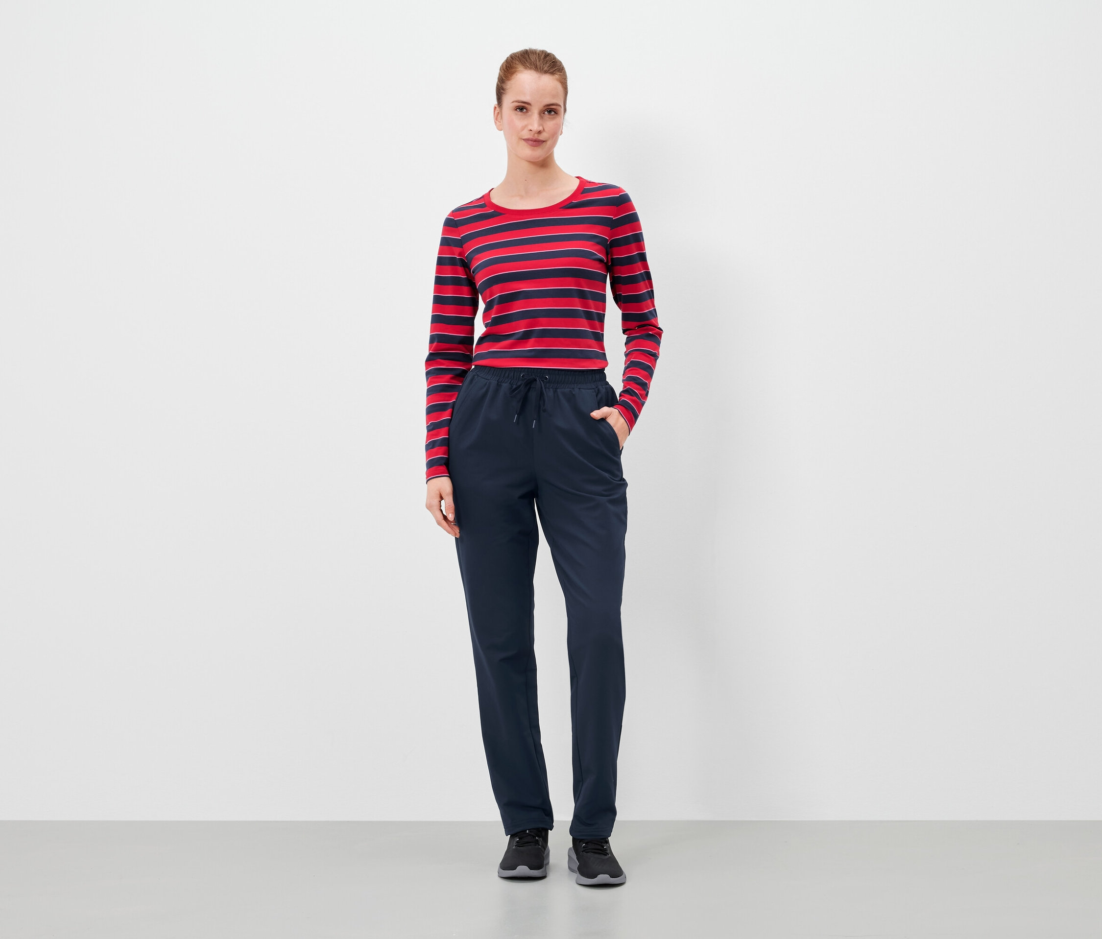 Kobieta stoi z rękami w kieszeniach granatowych spodni i ma na sobie czerwono-niebieską bluzkę w paski.