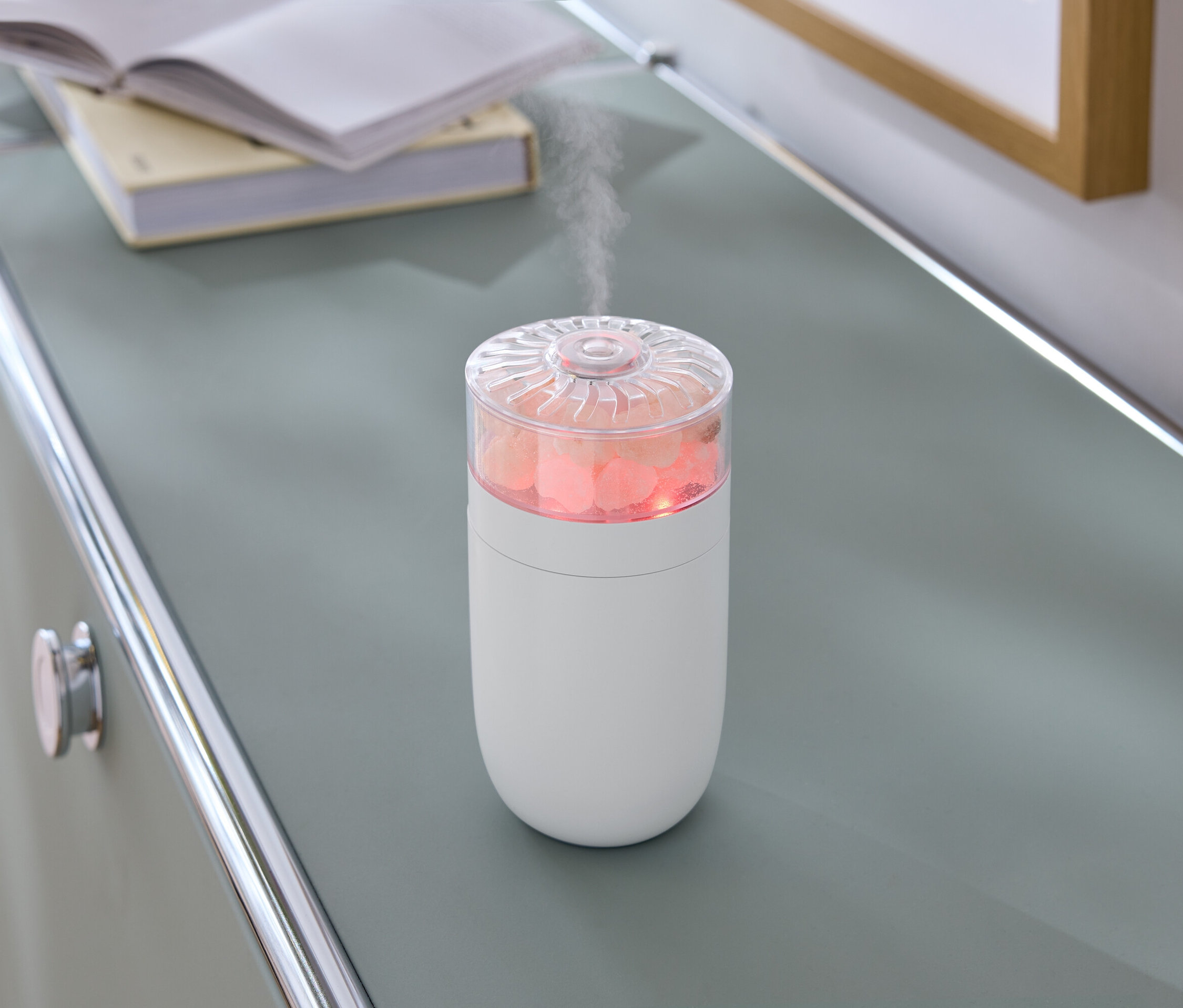 Na szarej komodzie stoi biały nawilżacz powietrza z różowymi kamieniami i parą.