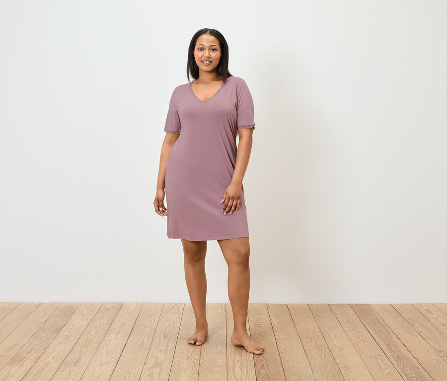 Kobieta stoi boso i ma na sobie krótką, fioletową koszulę nocną.