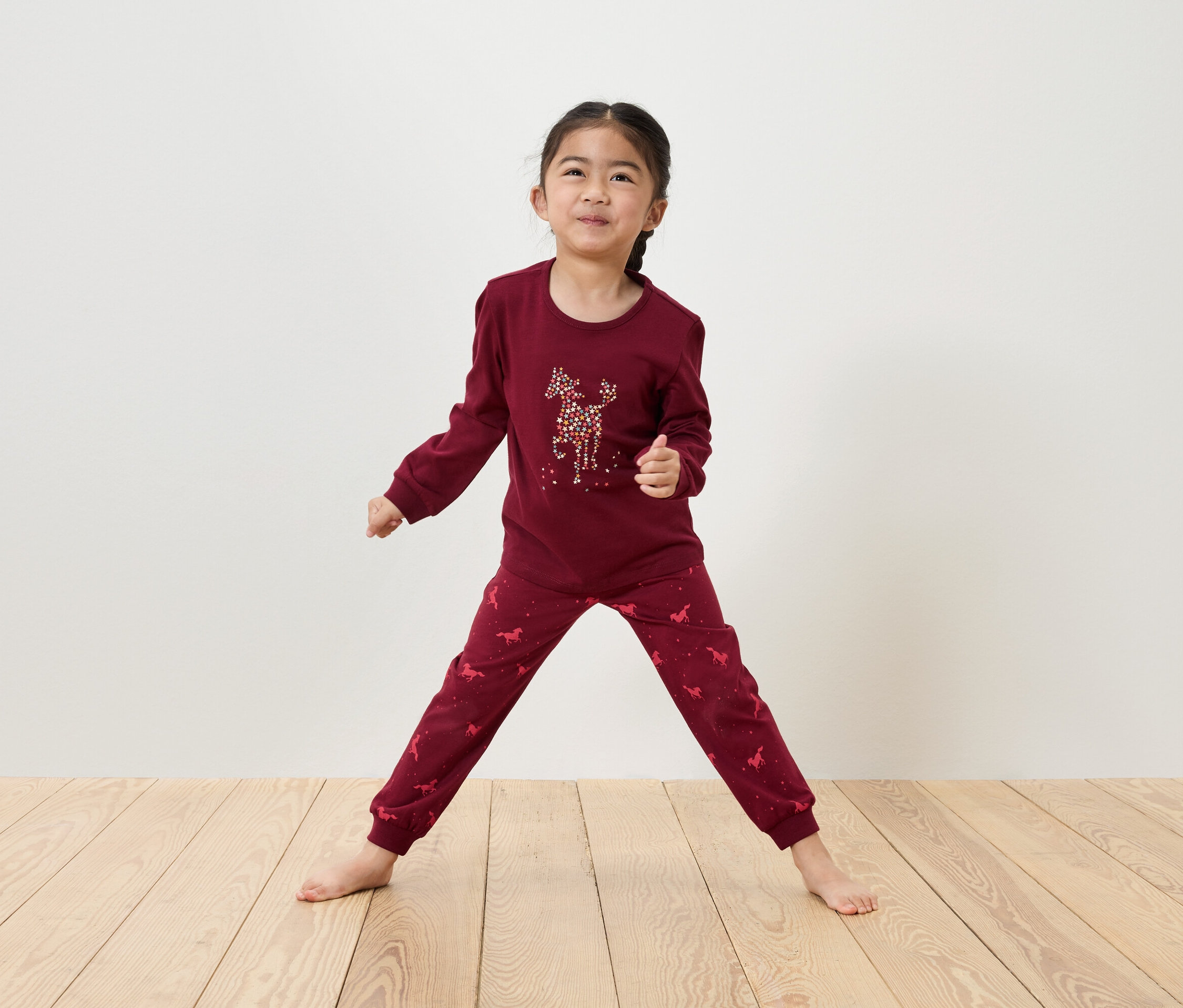 Dziewczynka w bordowej piżamie z motywem jelenia stoi na drewnianej podłodze.