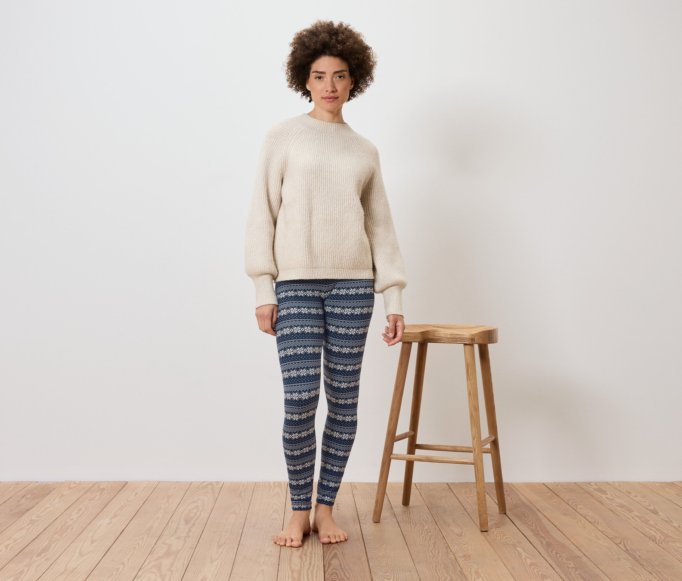 Kobieta stoi na tle białej ściany obok drewnianego stołka. Ma na sobie kremowy sweter i niebieskie legginsy we wzory.
