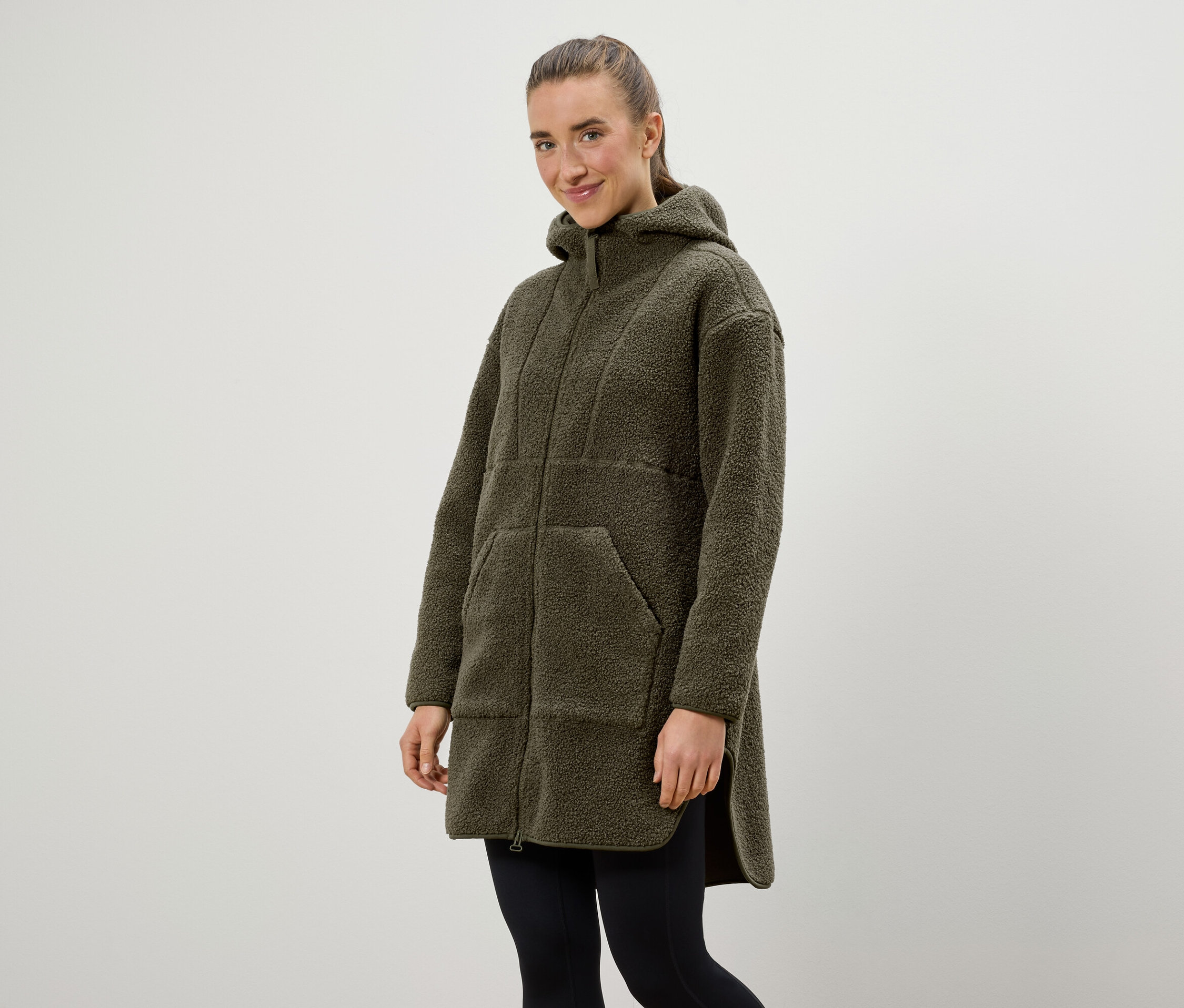 Kobieta ma na sobie oliwkowozielony płaszcz z kapturem i czarne legginsy.