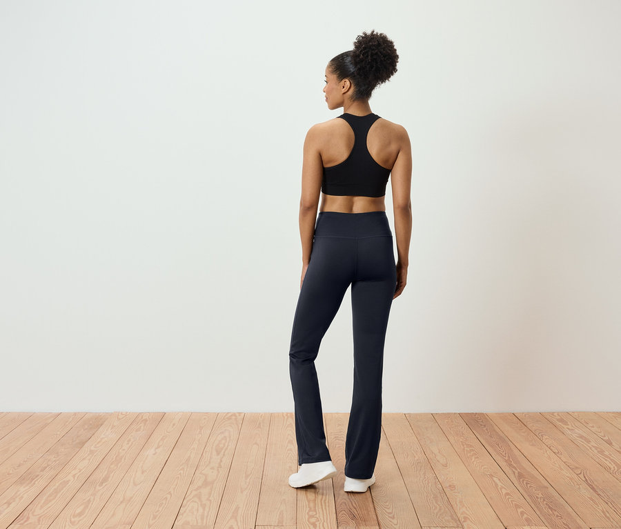 Widok z tyłu na kobietę z włosami upiętymi w kok, ubraną w czarny top sportowy i granatowe spodnie, stojącą na drewnianej podłodze.