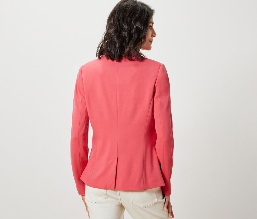 Kobieta stoi tyłem, ubrana w koralową marynarkę z dżerseju i jasne spodnie.