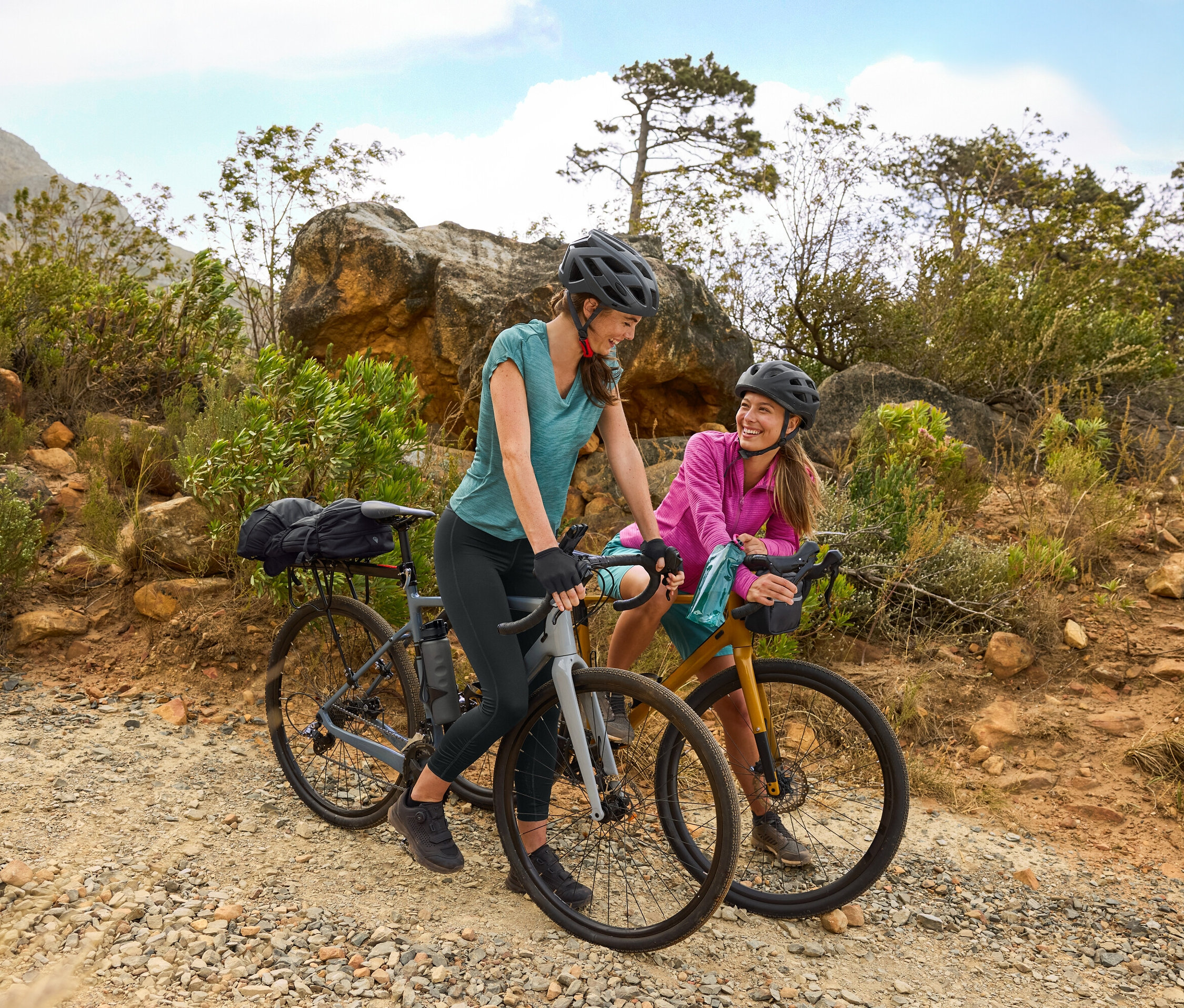 Dwie kobiety w kaskach stoją z rowerami na żwirowej drodze w pagórkowatym terenie. Bidon przymocowany jest do roweru.