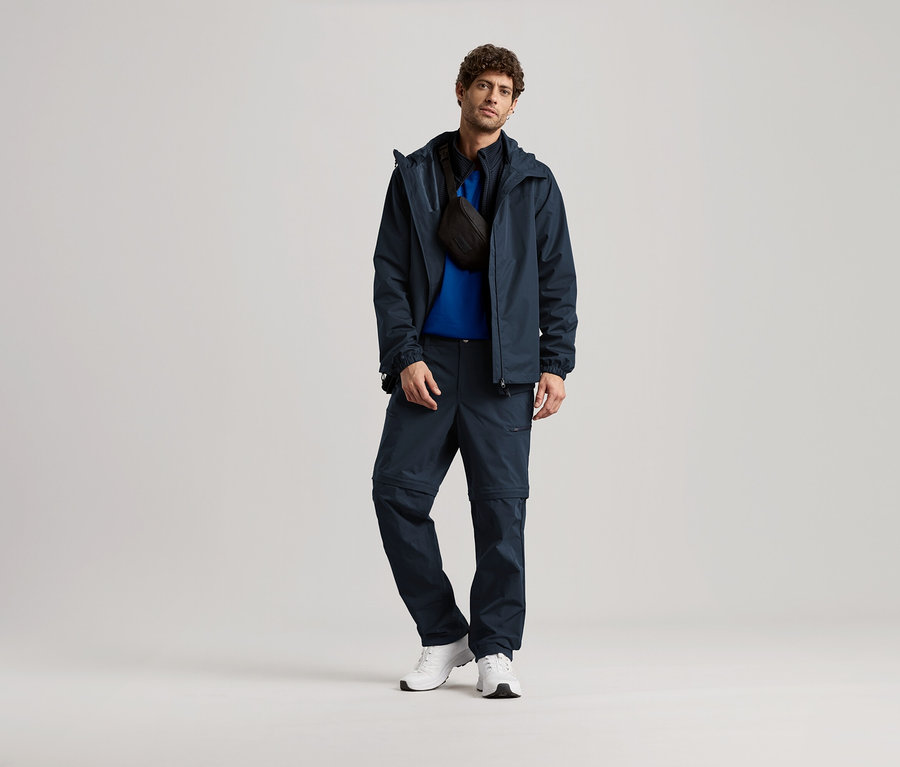 Mężczyzna stoi w granatowej kurtce, spodniach i białych tenisówkach, z czarną torbą na ramię.