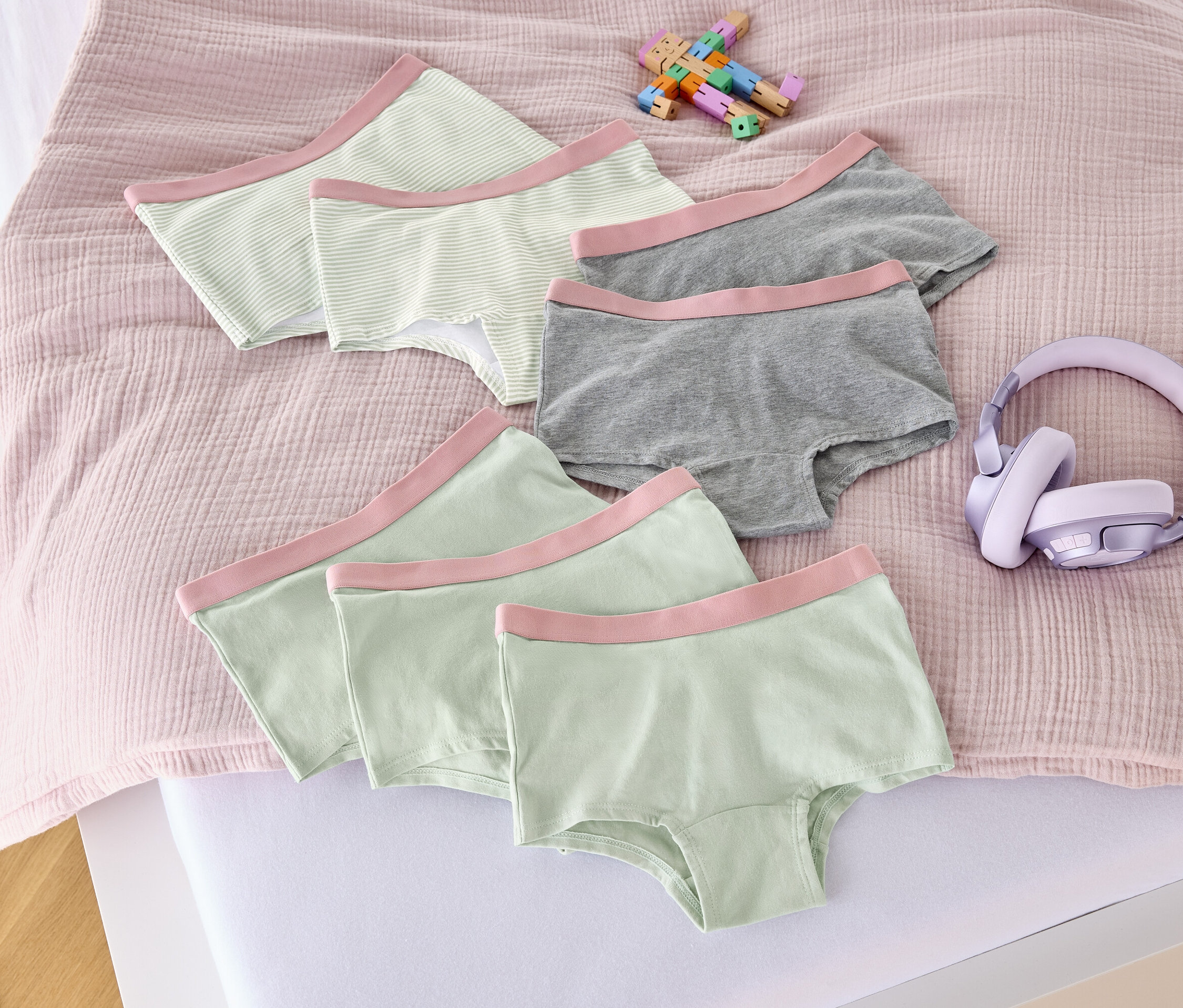 Na łóżku leży siedem majtek dziewczęcych w kolorze altrosa, zabawka i słuchawki.