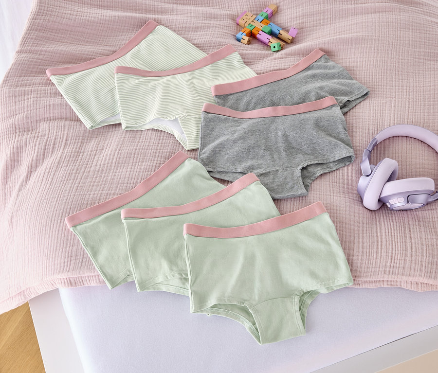 Na łóżku leży siedem majtek dziewczęcych w kolorze altrosa, zabawka i słuchawki.