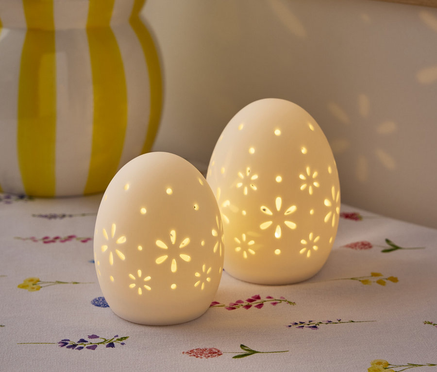 Dwie białe, jajowate lampy z kwiatowym wzorem stoją na stole z wazonem z kwiatami w tle.