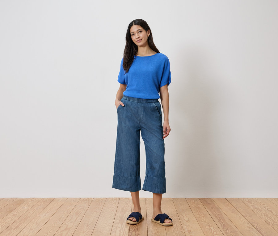 Kobieta stoi z ręką w kieszeni, ubrana w niebieski top, dżinsowe kuloty i sandały na tle białej ściany.