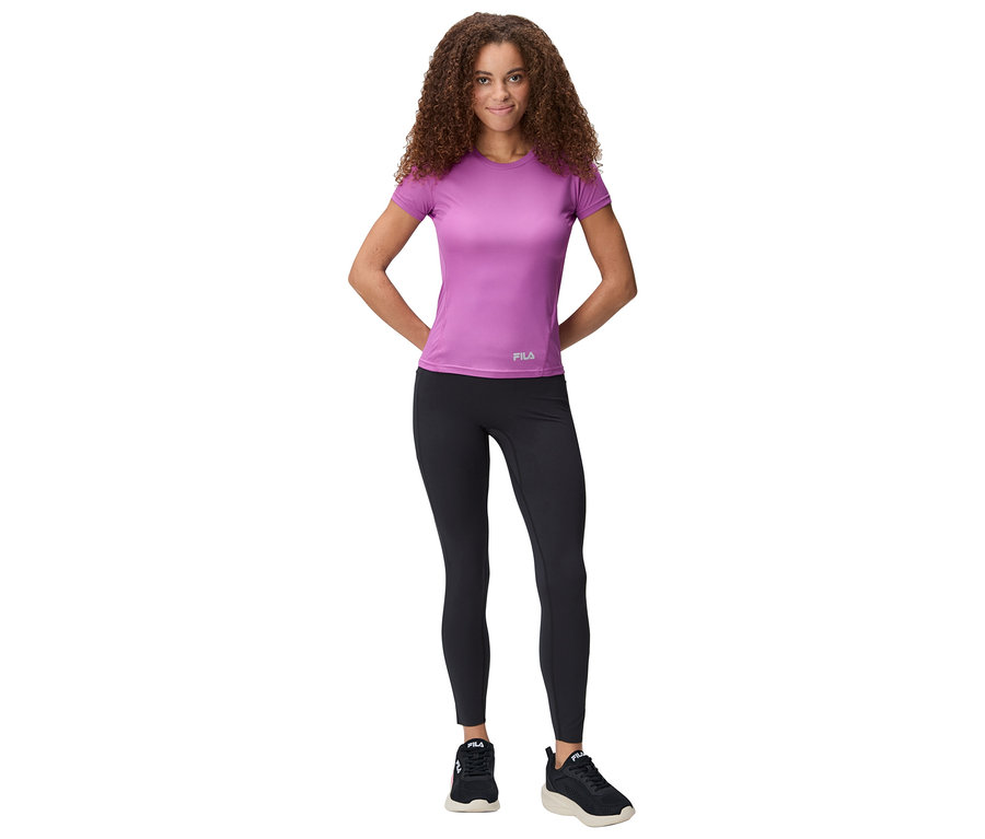 Kobieta stoi z rękami za plecami, ubrana w fioletową koszulkę, czarne legginsy i buty sportowe.