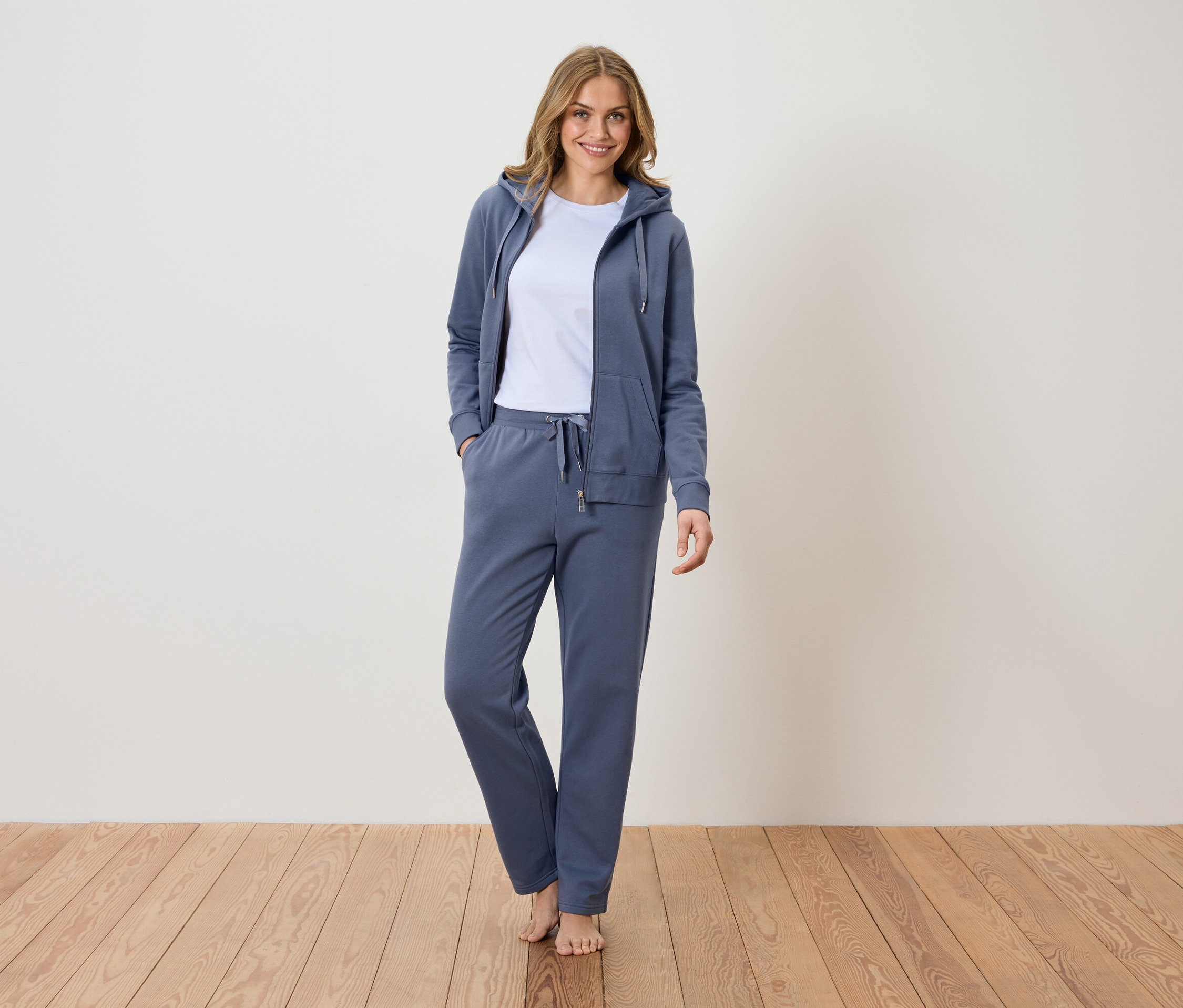 Kobieta stoi boso na drewnianej podłodze, ubrana w niebieski dres z bluzą z kapturem i białą koszulkę.