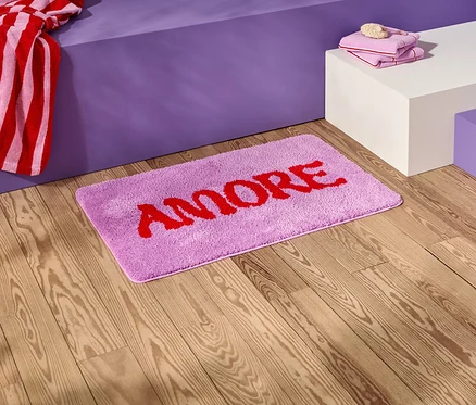 Na drewnianej podłodze leży liliowa mata łazienkowa z czerwonym napisem "AMORE".