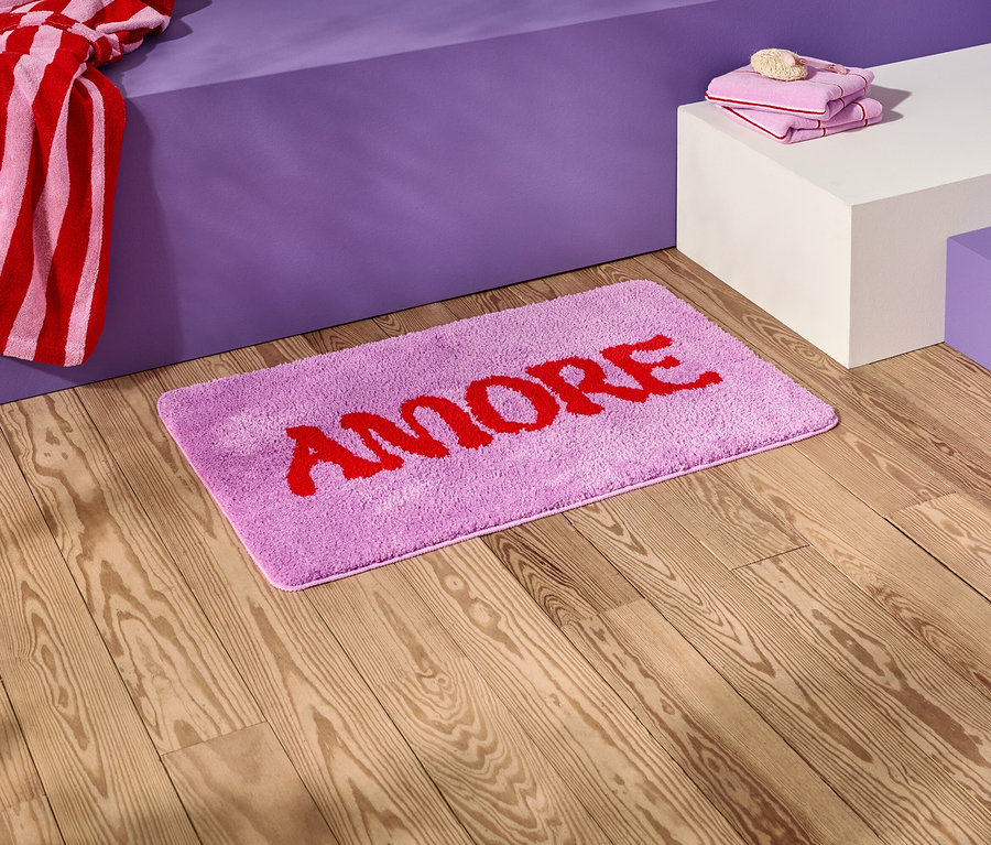 Na drewnianej podłodze leży liliowa mata łazienkowa z czerwonym napisem "AMORE".