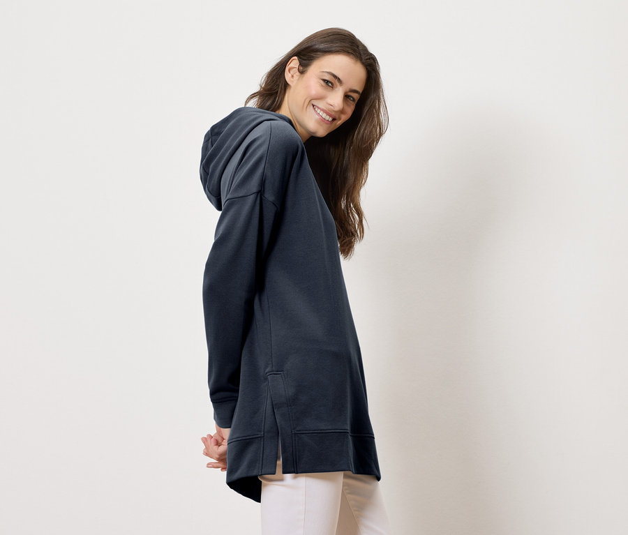 Kobieta w długiej, granatowej bluzie z kapturem i jasnych spodniach uśmiecha się do kamery.