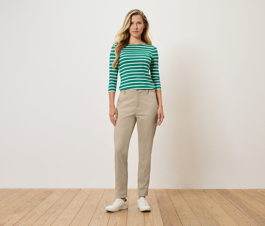 Kobieta stoi i ma na sobie zielono-białą bluzkę z długim rękawem w paski, beżowe spodnie i białe buty.