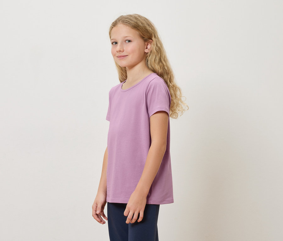 Dziewczyna z długimi blond włosami w fioletowej koszulce i granatowych spodniach stojąca na białym tle.