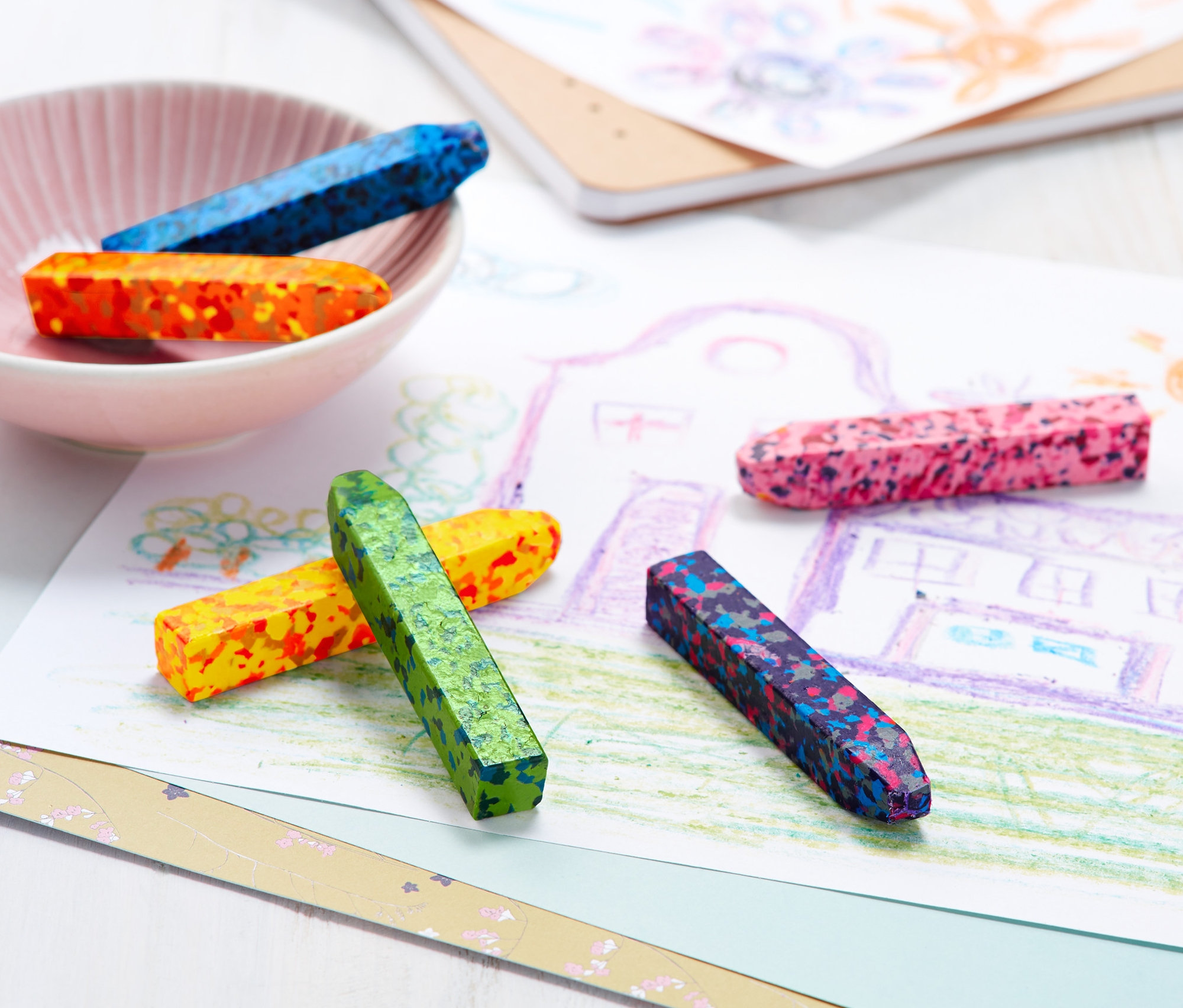 Na stole leży rysunek domu i słońca. Na rysunku leżą kolorowe Kredki świecowe konfetti, 6 sztuk . Dodatkowe kredki znajdują się w misce.