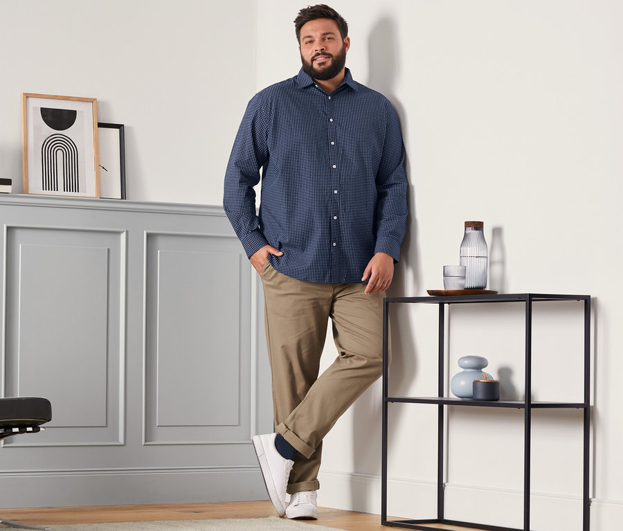 Mężczyzna w koszuli Comfort Fit, karmelowych spodniach chino i granatowych skarpetkach z wełny merino (4 pary, granatowe, czerwone i ciemnozielone) stoi w pokoju.