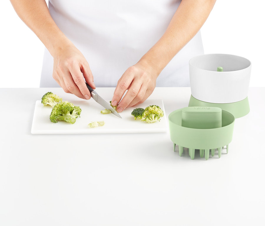 Osoba kroi brokuły na białej desce do krojenia obok rozdrabniacza do warzyw.