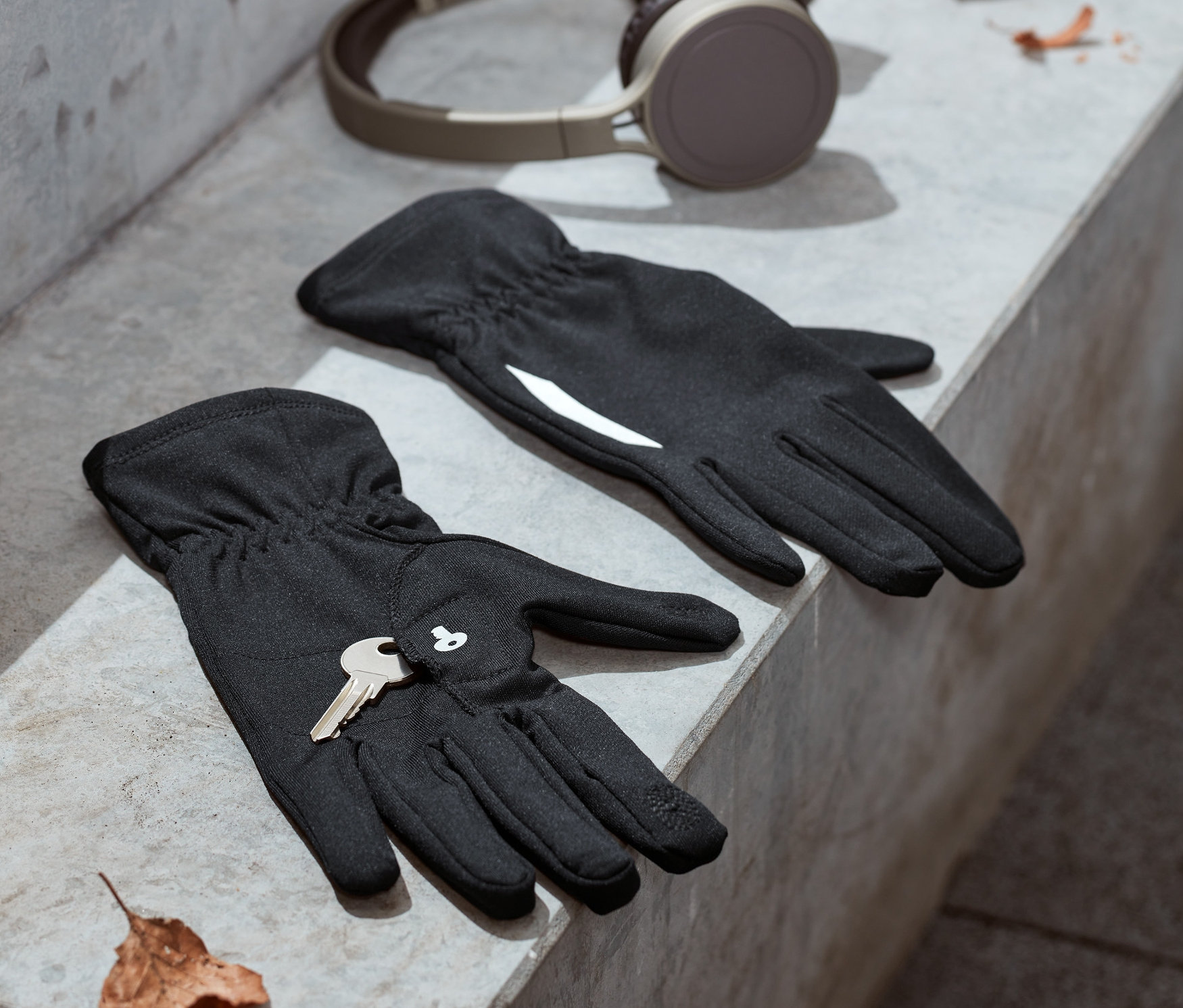 Na betonowych schodach leżą czarne rękawiczki do biegania z odblaskowym nadrukiem i klucz. W tle słuchawki i opadłe liście.