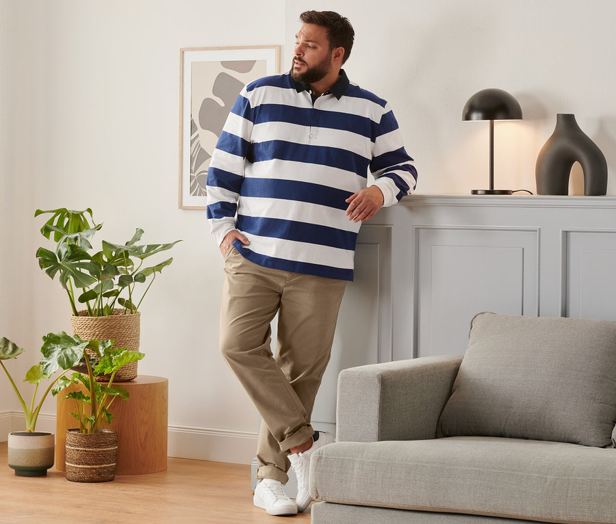 Mężczyzna opiera się o ścianę w karmelowych spodniach chino, niebiesko-białej koszulce polo z długim rękawem i butach typu sneaker.