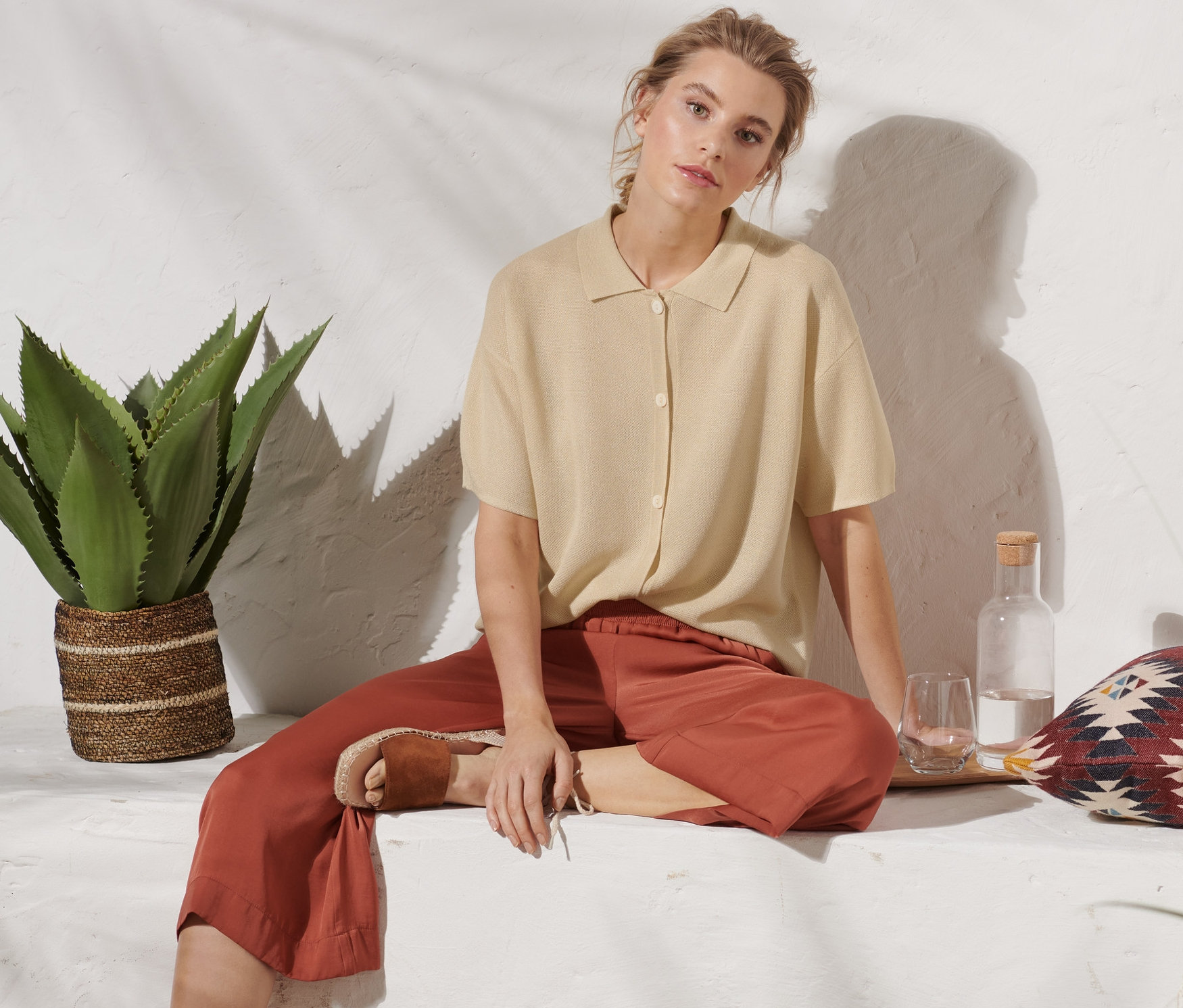 Kobieta siedzi i ma na sobie beżowy kardigan z kołnierzem, brązowe spodnie i sandały.