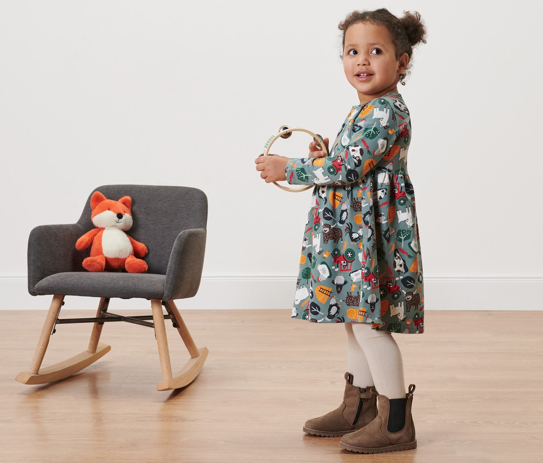 Dziewczynka w dżersejowej sukience niemowlęcej z nadrukiem trzyma tamburyn. Obok niej stoi szary fotel bujany z pluszowym lisem.