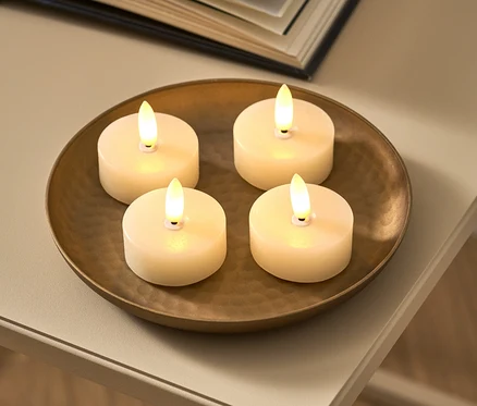 Na brązowej misce leżą cztery kremowe świeczki typu tealight LED z prawdziwego wosku. Obok leży otwarta książka.