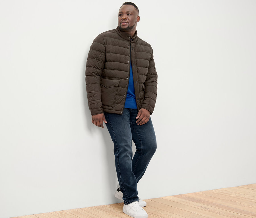 Mężczyzna oparty o ścianę ma na sobie kurtkę pikowaną, dżinsy i tenisówki.