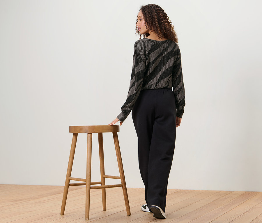 Kobieta stoi tyłem do aparatu, opierając się o drewniany stołek. Ma na sobie spodnie dresowe i bluzkę we wzór zwierzęcy.