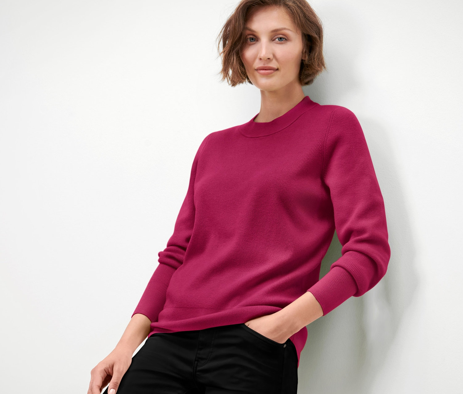 Kobieta opiera się o ścianę, ma na sobie sweter z delikatnej dzianiny w kolorze malinowej czerwieni i czarne spodnie.