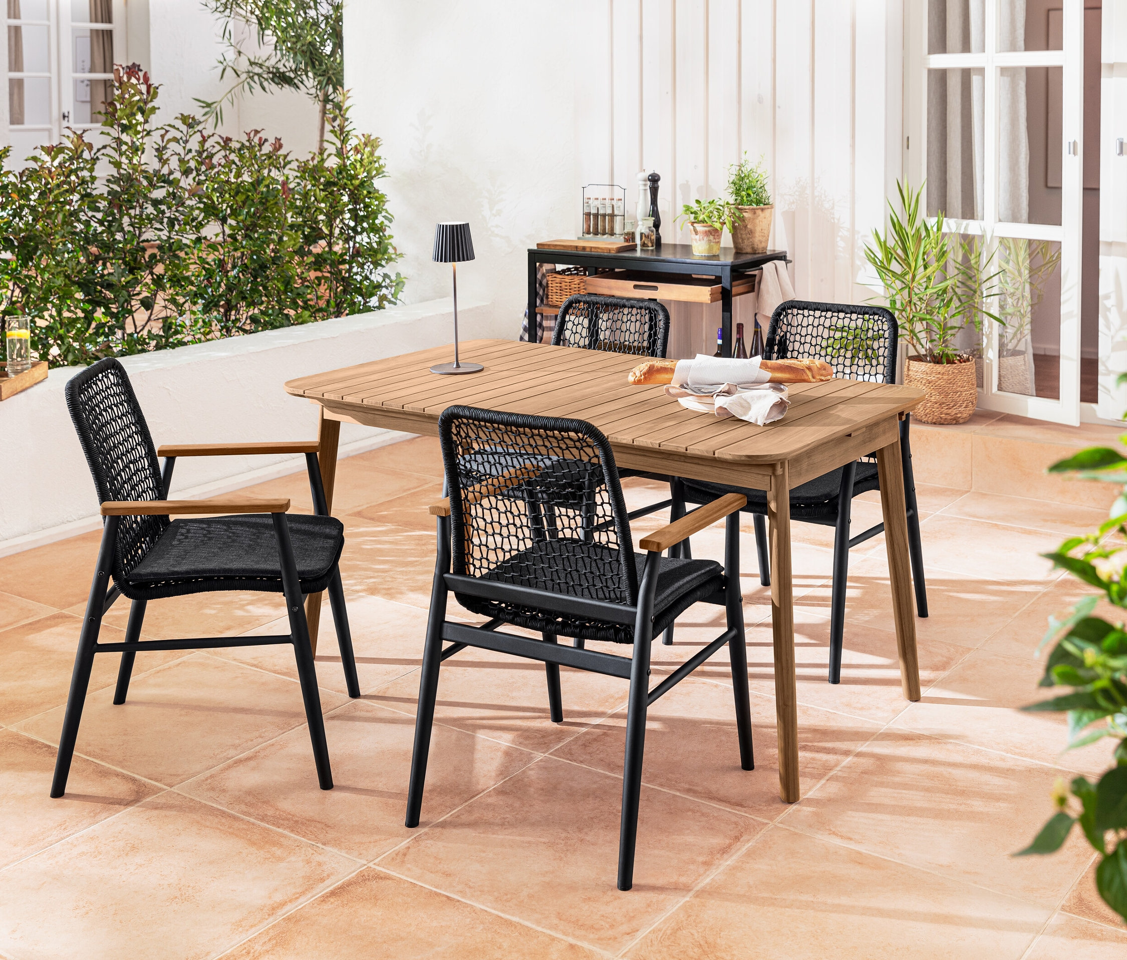 Cztery krzesła do jadalni z podłokietnikami z drewna tekowego i tekstylną plecionką stoją wokół rozkładanego stołu z drewna tekowego. Na stole stoi akumulatorowa lampa stołowa LED do użytku na zewnątrz, czarna.
