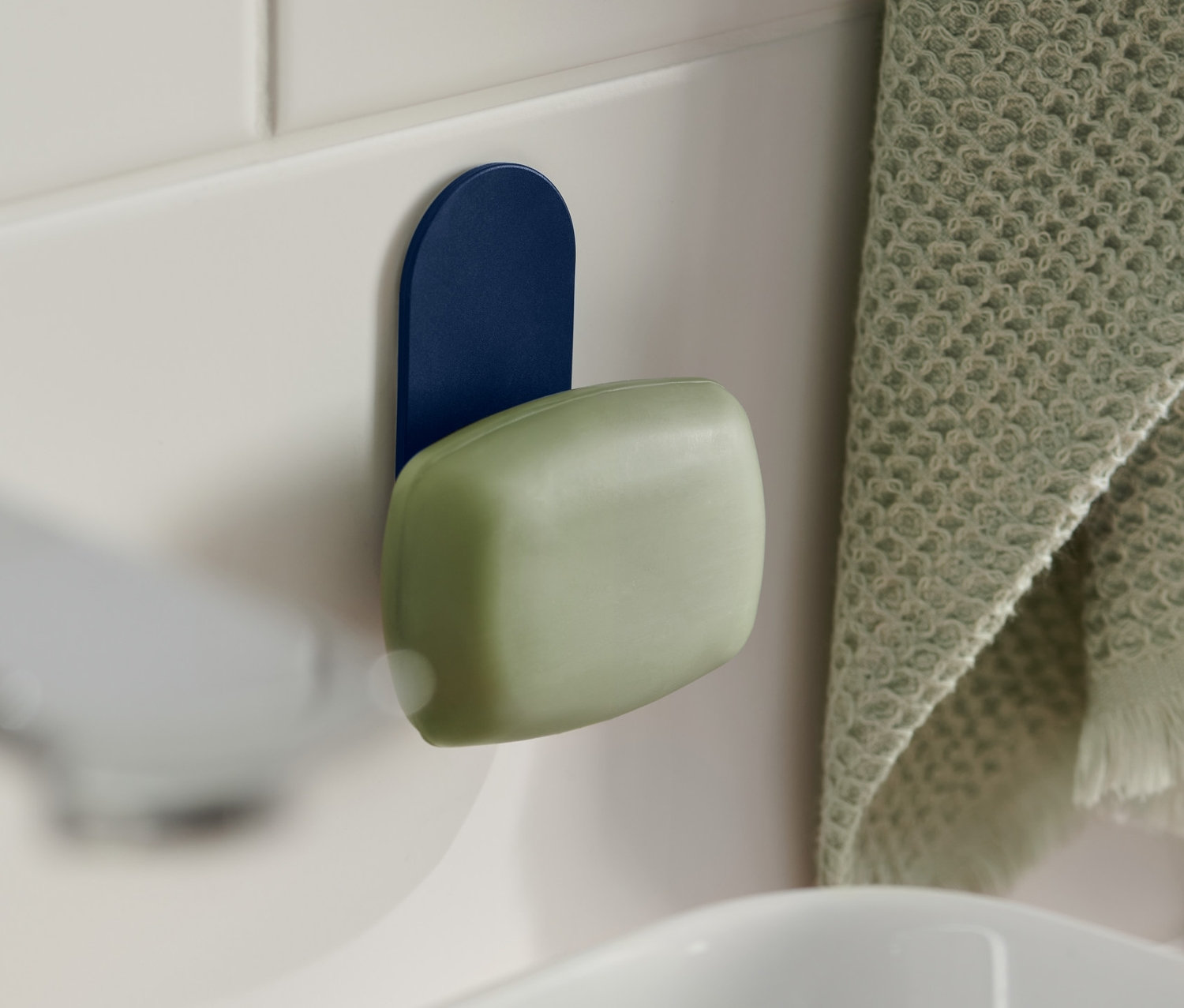 Zielone mydło wisi na magnetycznym uchwycie na mydło w kolorze niebieskim, przymocowanym do białych płytek obok umywalki. Po prawej stronie wisi zielony ręcznik.