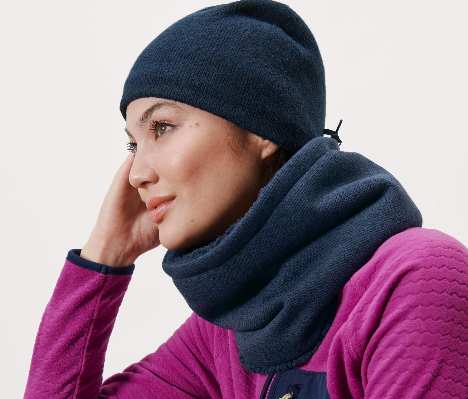 Kobieta oparta o drewnianą ławkę ma na sobie niebieski szalik z dzianiny polarowej i niebieską czapkę.